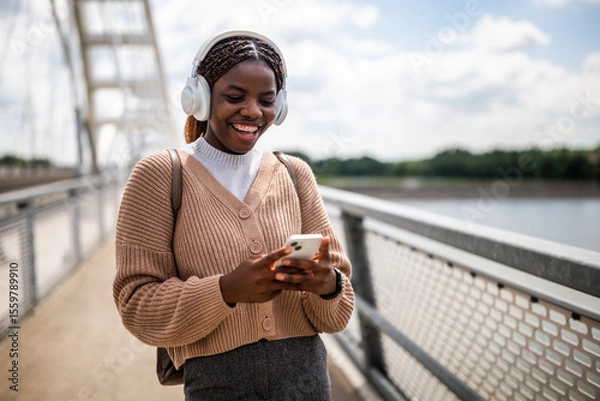 Fototapeta Happy young black woman is typing on her phone and smiling. She is enjoying sunny day outdoor and listening music while using smartphone.