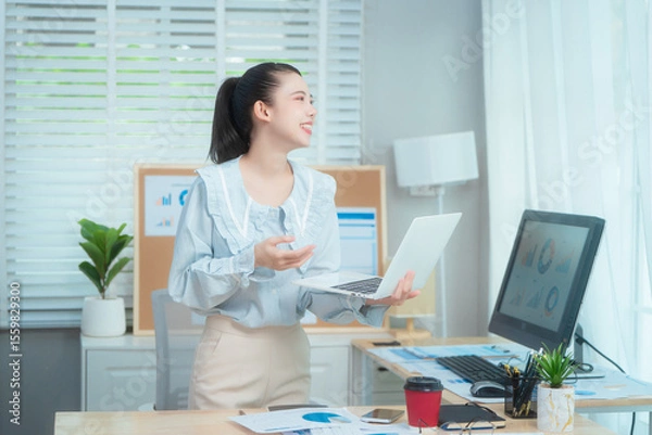 Fototapeta Confident beauty Asian office woman in a formal blouse hard working at her desk, reviewing a document with focus and a pen in hand. The workspace includes charts, a laptop, and a desktop computer.