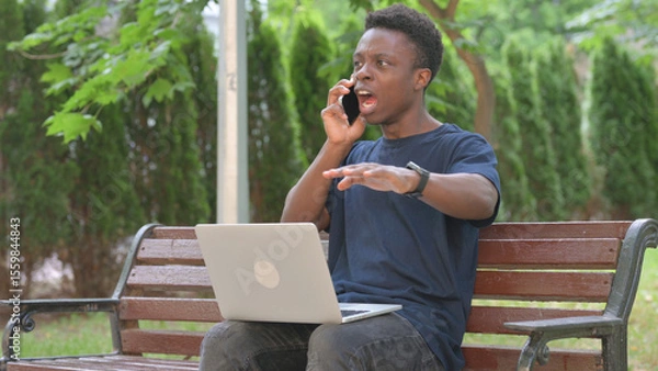 Fototapeta Angry African Man Talking on Phone While Using Laptop in Park
