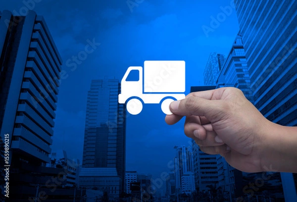 Fototapeta Hand holding delivery truck flat icon in fingers over modern office city tower and skyscraper, Business transportation service concept