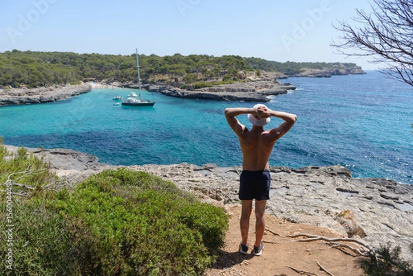 Obraz young athletic man looking from the shore at a sailing yacht at anchor in a beautiful sea bay, summer vacation