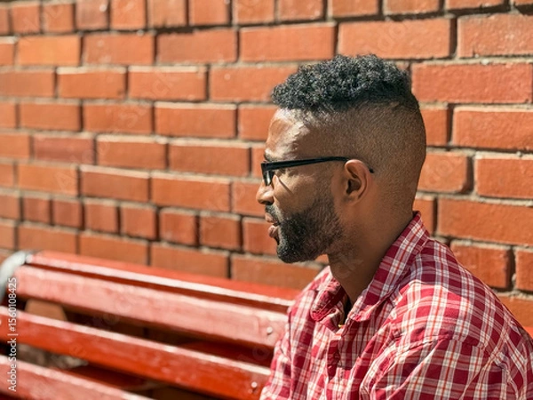 Obraz Portrait of a young man of African-American nationality wearing glasses with a beard sitting on a bench. A black guy in a plaid shirt on the background of a brick wall. Portrait of a black man. 