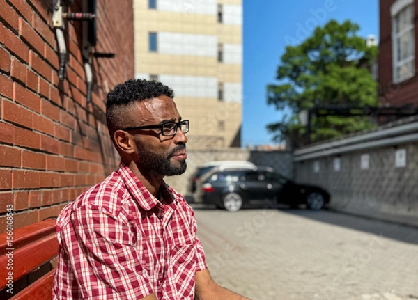 Obraz Portrait of a young man of African-American nationality wearing glasses with a beard sitting on a bench. A black guy in a plaid shirt on the background of a brick wall. Portrait of a black man. 