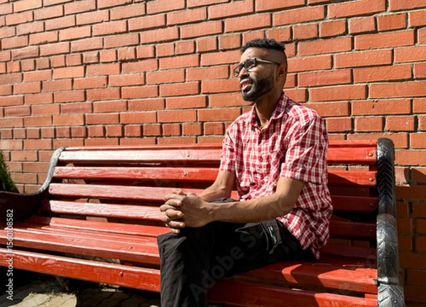 Obraz Portrait of a young man of African-American nationality wearing glasses with a beard sitting on a bench. A black guy in a plaid shirt on the background of a brick wall. Portrait of a black man. 