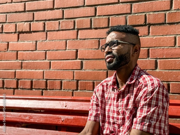 Obraz Portrait of a young man of African-American nationality wearing glasses with a beard sitting on a bench. A black guy in a plaid shirt on the background of a brick wall. Portrait of a black man. 
