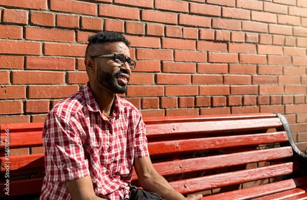 Obraz Portrait of a young man of African-American nationality wearing glasses with a beard sitting on a bench. A black guy in a plaid shirt on the background of a brick wall. Portrait of a black man. 