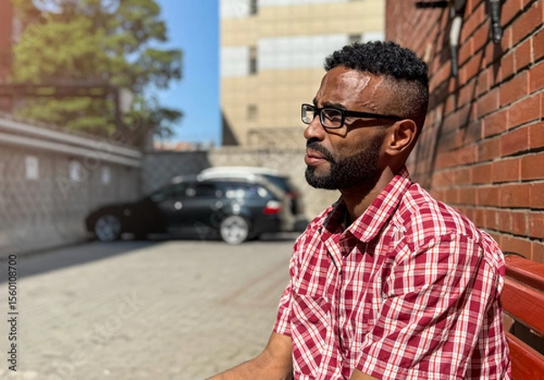 Obraz Portrait of a young man of African-American nationality wearing glasses with a beard sitting on a bench. A black guy in a plaid shirt on the background of a brick wall. Portrait of a black man. 