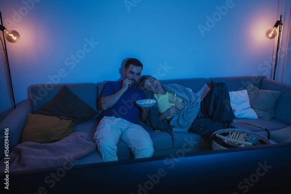 Obraz Couple watching TV at night  while sharing a bowl of popcorn and wine on sofa