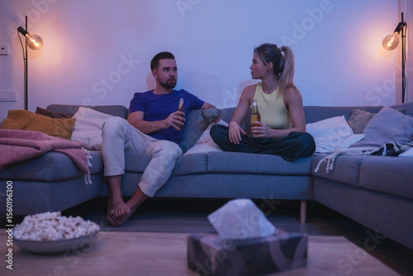 Obraz Couple sitting on a couch, enjoying beer and nachos in a cozy living room
