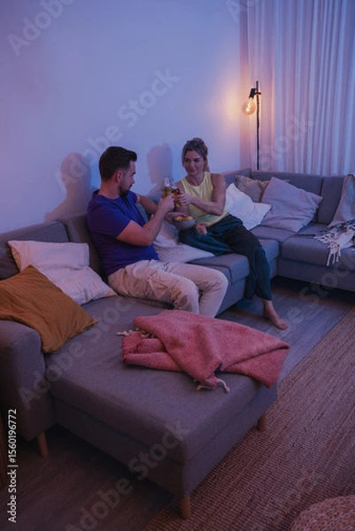 Obraz Couple sitting on a couch, enjoying beer and nachos in a cozy living room