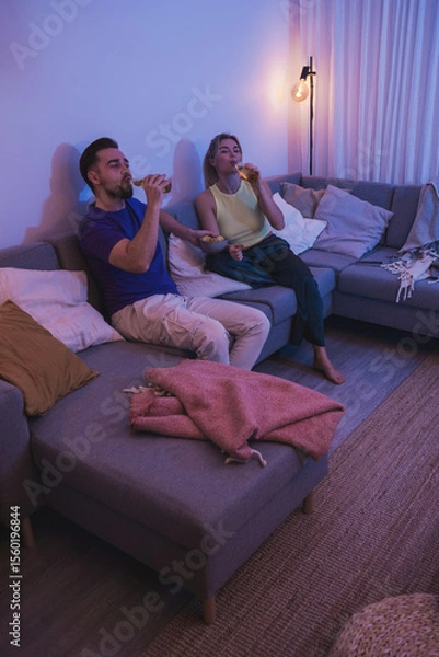 Obraz Couple sitting on a couch, enjoying beer and nachos in a cozy living room