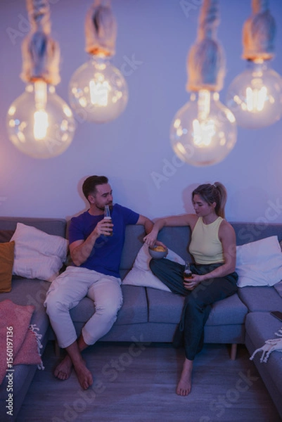 Obraz Couple sitting on a couch, enjoying beer and nachos in a cozy living room