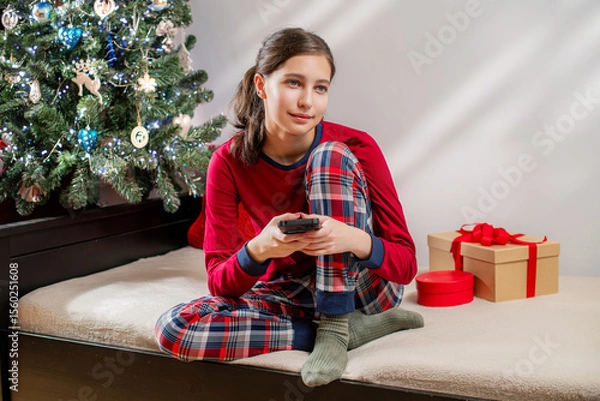 Fototapeta Smiling girl in red pajamas using mobile phone near Christmas tree