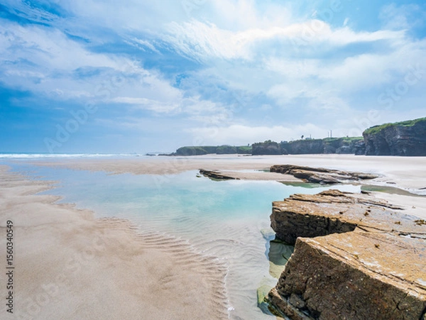 Fototapeta Low tide at As Catedrais beach Galicia Spain reveals sandstone rocks tidal pools reflecting sky cliffs lining wide shore under bright light peaceful calm natural wonder