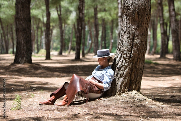 Fototapeta A young, pretty woman dressed in riding breeches and riding clothes and a typical Spanish hat rests seated in the shade of a pine tree on a pilgrimage day in Spain.