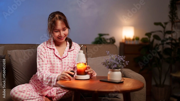 Fototapeta A contented Asian adult female enhances her restful evening with an aromatic candle for a sense of calm and wellness at home