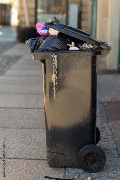 Fototapeta Overflowing trash can on a quiet city street showcases the need for better waste management and environmental awareness at the start of a sunny day