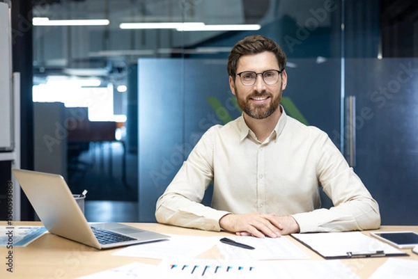 Fototapeta Portrait of a young smiling man in a shirt and glasses sitting in the office at a table with a laptop and documents, looking at the camera