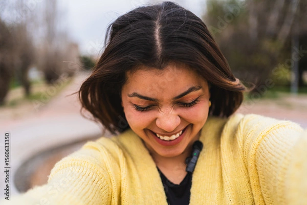 Fototapeta Young female influencer wearing a yellow sweater is happily laughing while recording a video blog in a park