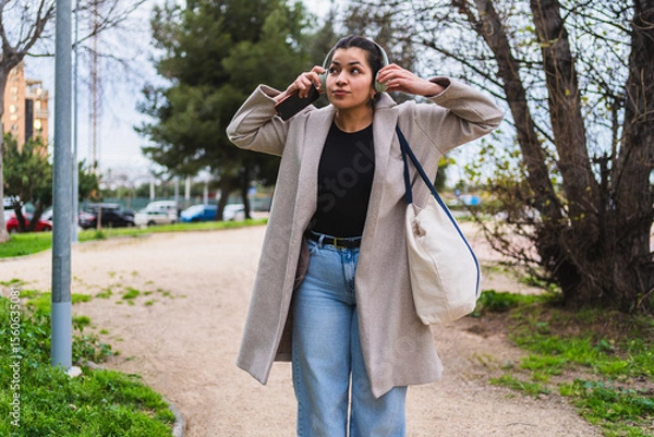 Fototapeta Young stylish influencer adjusting headphones while listening to music on her smartphone and walking in a park