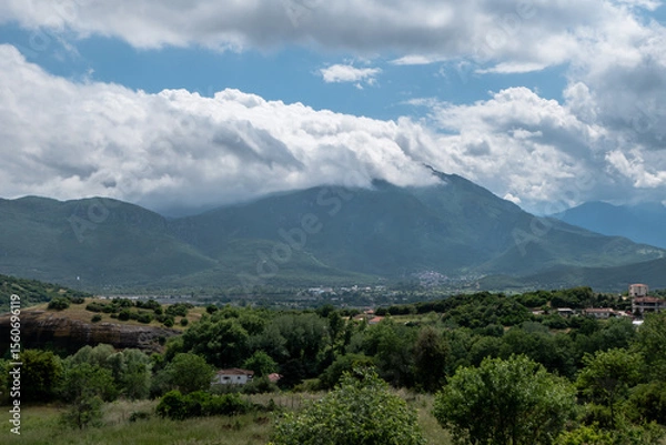 Fototapeta A serene panoramic view of lush green hills and a rural village nestled in a valley with dramatic clouds rolling over a mountain range in the background.