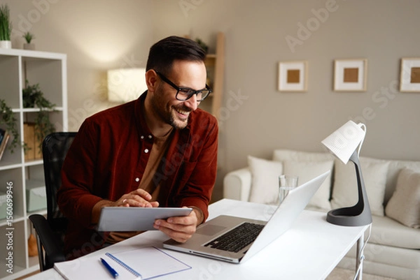 Obraz Freelancer smiling and working from home using laptop and tablet