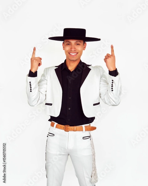 Fototapeta Young chilean Huaso wearing traditional white suit with black details, pointing upwards with index fingers, while smiling