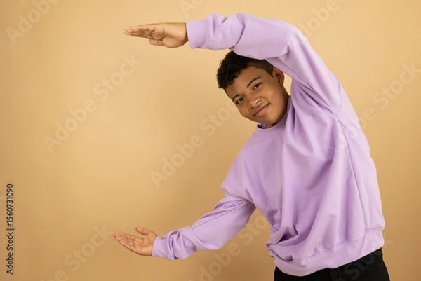 Fototapeta Brown skinned Latino teenager wearing a lilac sweatshirt pointing with both hands at a copy space on a light brown background, advertising space to promote