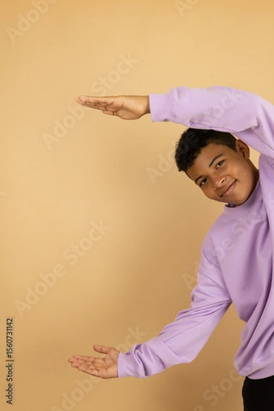 Fototapeta Vertical image of a young brown skinned Latino man wearing a lilac sweatshirt pointing with both hands at a copy space on a light brown background, advertising space to promote