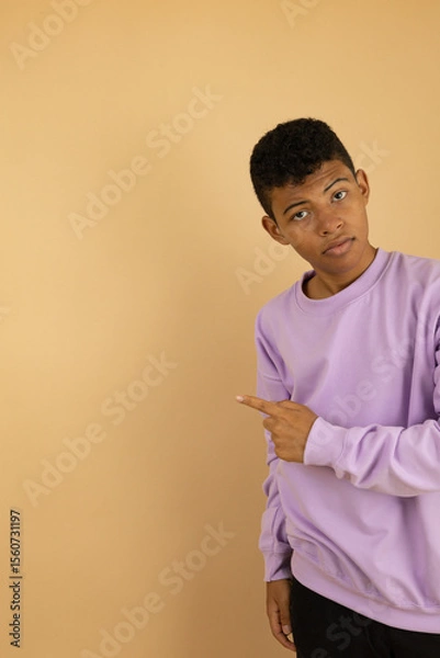Fototapeta Vertical image of a young brown skinned Latino man wearing a lilac sweatshirt pointing with his fingers at a copy space on a light brown background, advertising space to promote