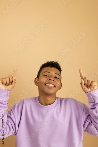 Fototapeta Cheerful brown skinned Latino teenager wearing a purple sweatshirt pointing with his index fingers at a top with copy space, Young man highlighting a blank space