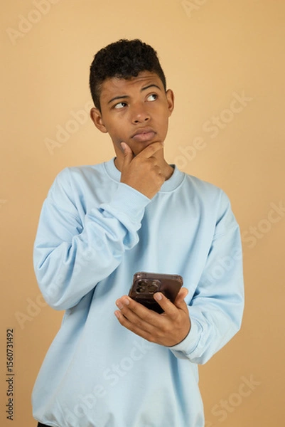 Fototapeta Vertical image of a dark skinned Latino teenager wearing a pastel blue sweatshirt with a thoughtful expression while holding his cell phone on a light brown background with copy space