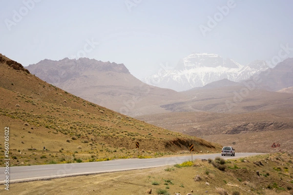 Fototapeta A car winds along a remote road through Yazd’s arid terrain, with dramatic snow-capped mountains rising in the background beneath a sunlit sky.