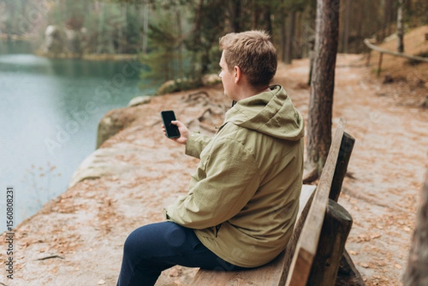 Obraz Lonely 30s blonde man with phone is sitting on the bench of a forest lake. Autumn landscape, calmness, tranquility, travel concept. A man on the bank of a lake is enjoying the nature.