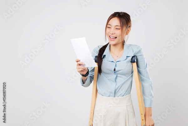 Fototapeta Cheerful woman with leg injury holds bill payment paper and smiles, using crutches for support and crutches to balance during recovery. isolated studio white background
