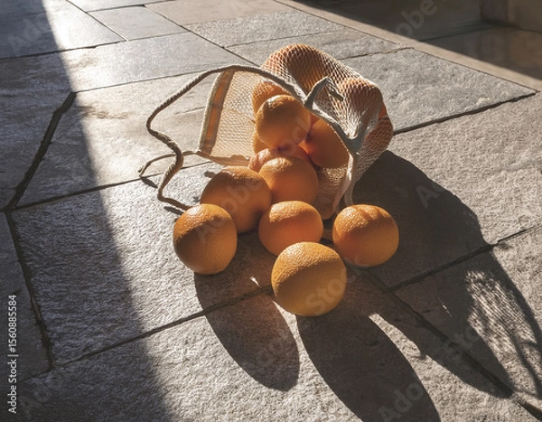 Fototapeta A group of oranges spilled from a market bag onto a sunlit stone floor with rich shadows
