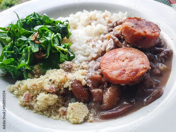 Fototapeta Brazilian food, with white plate with rice, feijoada, sautéed kale and farofa and blurred background