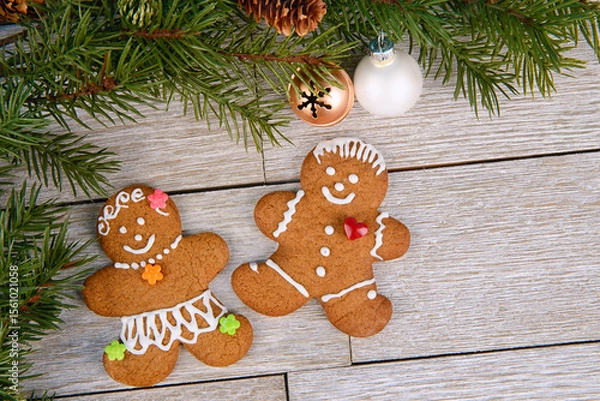 Obraz Gingerbread man and woman cookies with icing on rustic wooden table, pine branches and baubles create a cozy festive scene, perfect for holiday ads, cards, and winter celebration concepts.