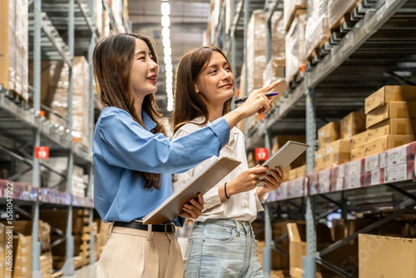 Obraz Two young asian women share insights and strategies while inspecting numerous packages and boxes engage in lively discussion while navigating a bustling warehouse filled with packages