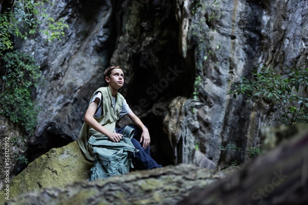 Obraz A hiker rests on a mossy rock inside a rocky canyon, surrounded by lush greenery. The peaceful scene captures a moment of solitude, reflection, and connection with nature during an outdoor adventure.