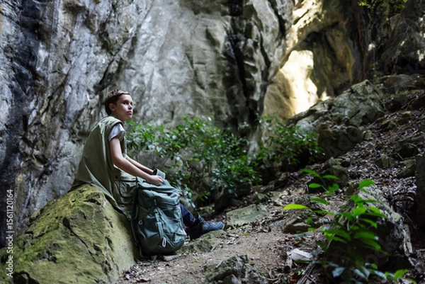 Obraz A hiker rests on a mossy rock inside a rocky canyon, surrounded by lush greenery. The peaceful scene captures a moment of solitude, reflection, and connection with nature during an outdoor adventure.