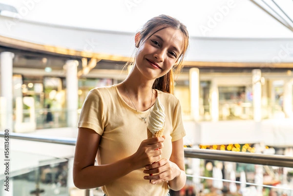 Obraz Joy radiates from a young asian woman savoring a delicious ice cream cone while standing in a bustling shopping center. The sun illuminates her smile as she enjoys this sweet treat.