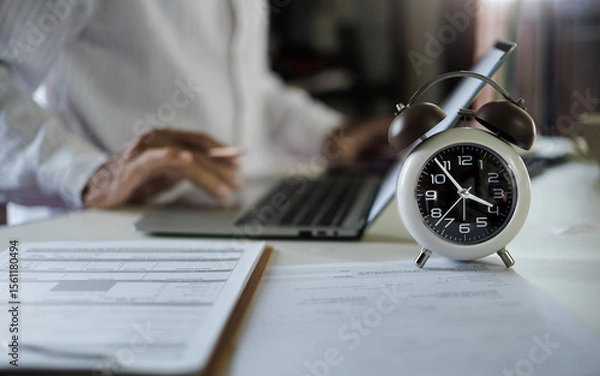 Fototapeta selective focus on alarm clock, asian businessman working with laptop and document on table before work time end in a day,  time management concept, routine work, office hour