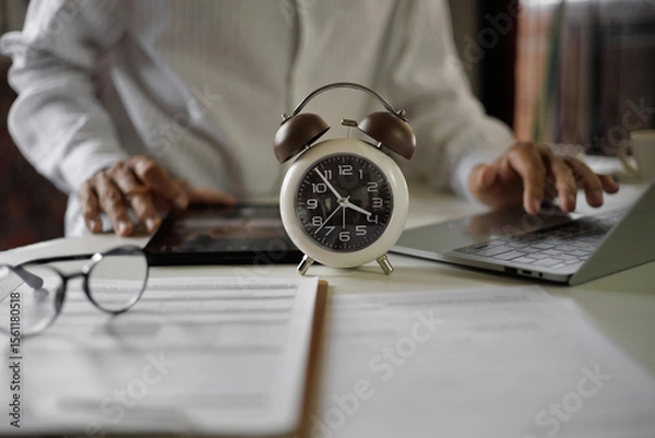 Fototapeta selective focus on alarm clock, asian businessman using laptop and tablet working on table before work time end in a day,  time management concept, routine work, office hour, blur background