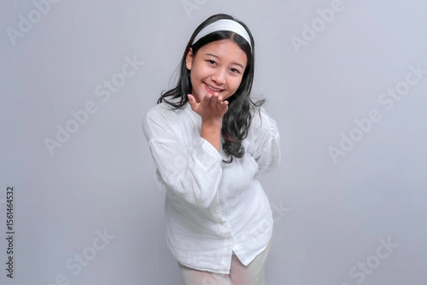 Fototapeta Portrait of a cheerful Asian woman in a white outfit smiling and blowing a kiss toward the camera with one hand, conveying affection, happiness, and positive emotion in studio.