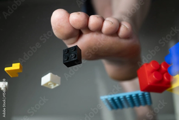 Obraz A foot is shown stepping on colorful construction toy bricks scattered on the floor, highlighting the chaos of playtime. The setting suggests a fun but messy environment created by children.