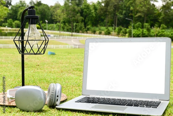 Obraz A laptop is open on a grassy field with a lamp and headphones nearby. The scene suggests a relaxed and leisurely atmosphere, possibly for someone to work or study outdoors