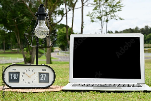 Obraz A white laptop with a black screen is placed on the green grass, with a lamp and a clock beside it.