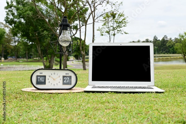 Obraz A white laptop with a black screen is placed on the green grass, with a lamp and a clock beside it.