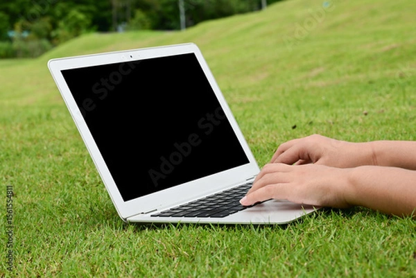 Obraz A person is typing on a laptop in a grassy field. The laptop is open and the screen is black